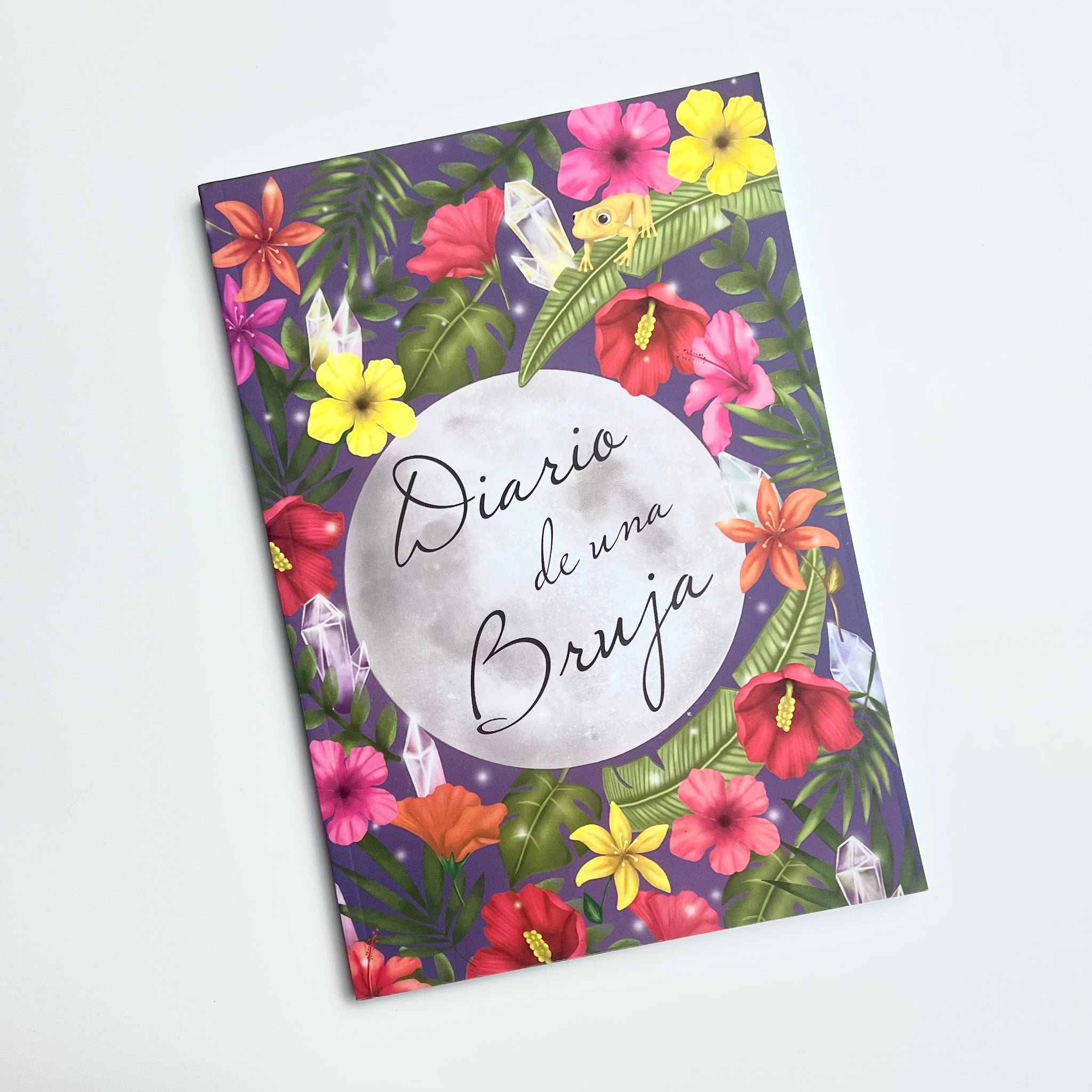 Softcover journal. On the cover it has an illustration that shows a full moon. On the moon is written the text Diario de una bruja or Diary of a witch. The moon is surrounded by tropical flowers, crystals and a coquí, a tiny Puerto Rican frog. 