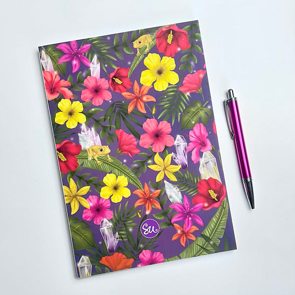 Image shows backside of the Softcover journal. The backcover shows an illustration of tropical flowers, crystals and the coquí, a tiny Puerto Rican frog on top of some leaves. Besides the journal there is a pink pen to compare sizes.